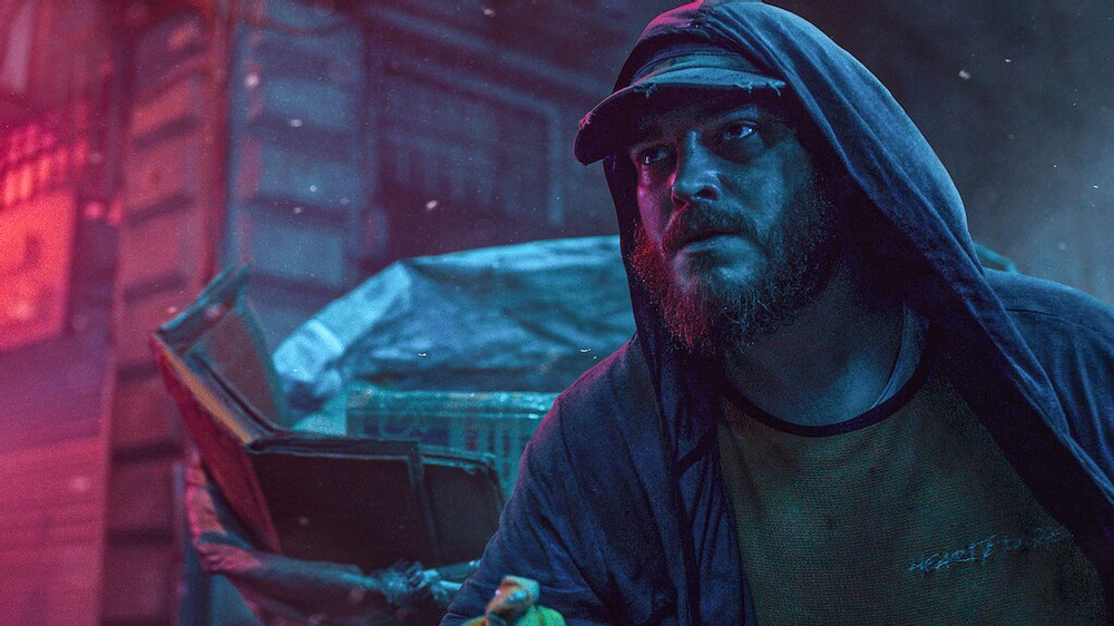 A man stands, illuminated in pink, red and blue lighting the street, with a cap on his head and a hoodie pulled over him as well. He looks off into the distance (left) and a sack of different waste materials, like cardboard, can be seen behind him where he looks. He also has a beard and moustache; looks quite tired and glistens with rain as well as sweat from working. 